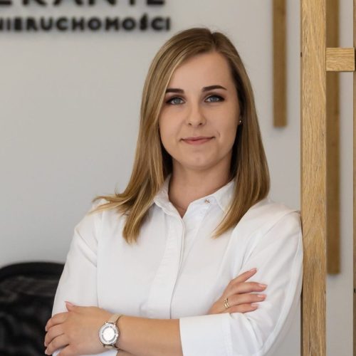 ED4A4766 Kobieta w białej koszuli stojąca z założonymi rękami, z lekkim uśmiechem, na tle biura nieruchomości.